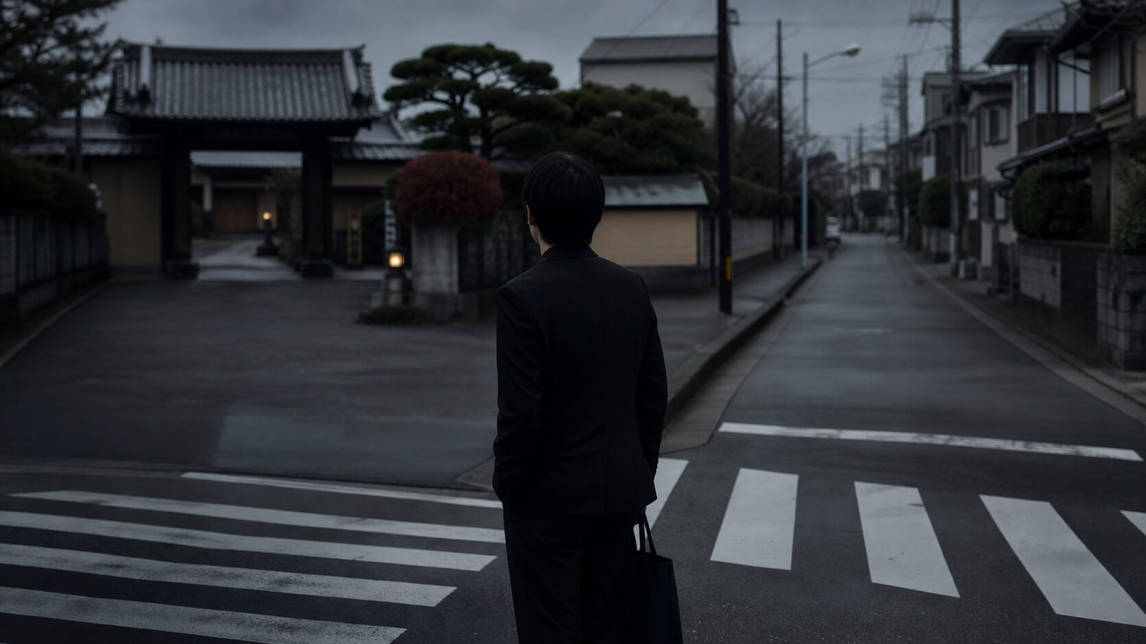 葬儀会場へ続く道と住宅街の分岐に立つ黒い礼服の人物